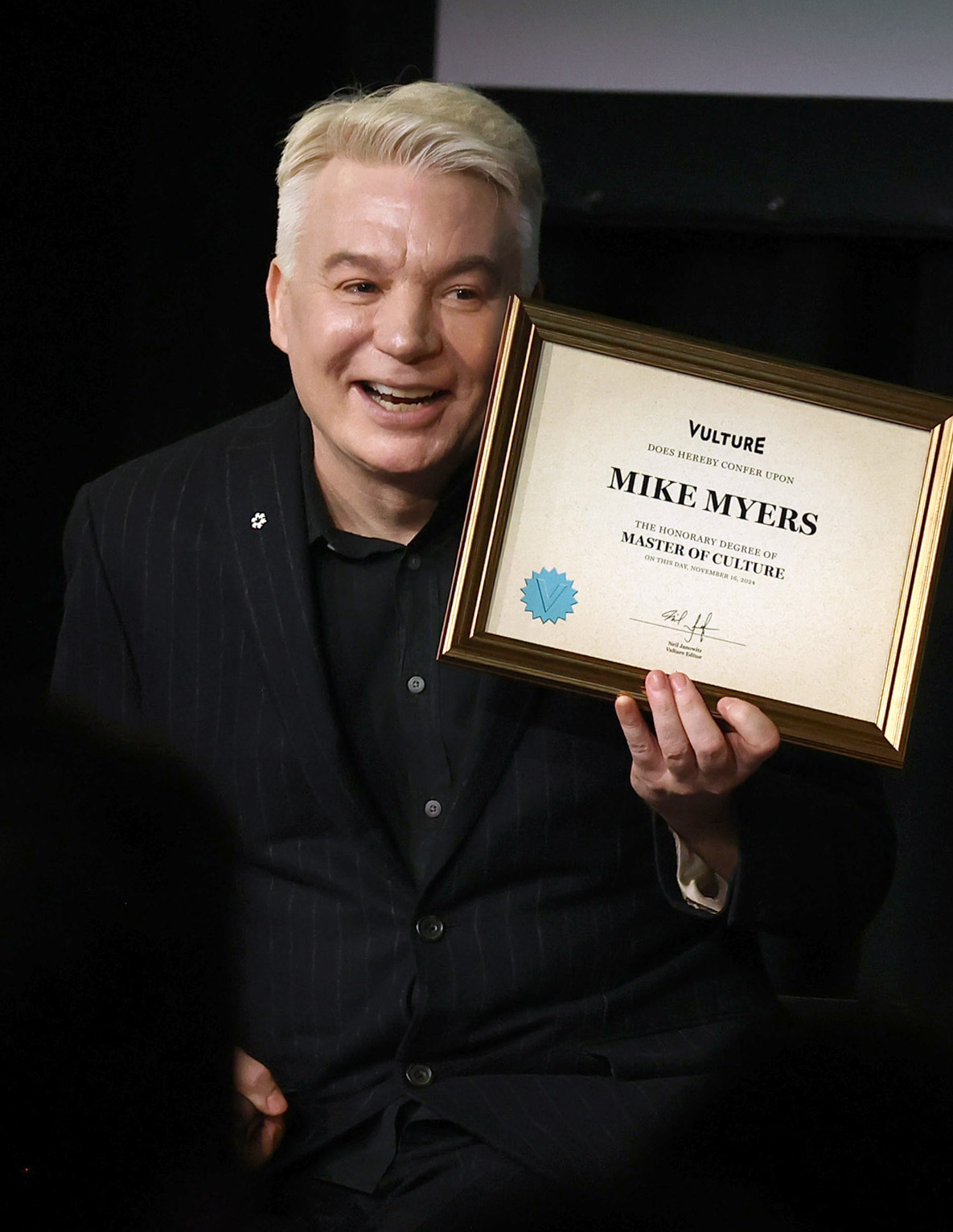 Mike Myers smiling while holding a framed certificate titled "Master of Culture" from Vulture. He is dressed in a black suit with subtle pinstripes.